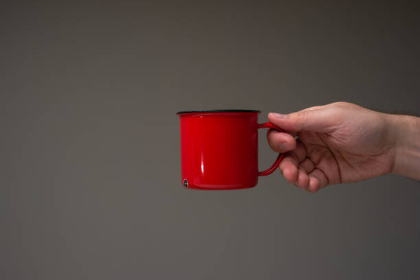 Red enameled tin camping cup or mug held in hand by a Caucasian male. Close up studio shot, isolated on gray background stock photo