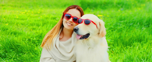 Portrait of happy woman with her Golden Retriever dog wearing sunglasses on green grass in summer park