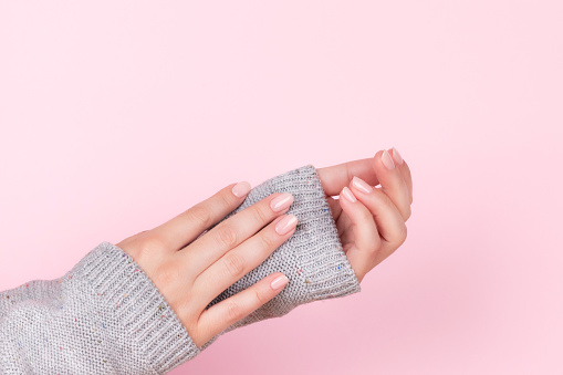 Female hands in gray knitted sweater with beautiful natural manicure - pink nude nails on pink background. Nail care concept