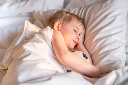 Pretty toddler boy sleeping with a soccer ball on a white bed linen.