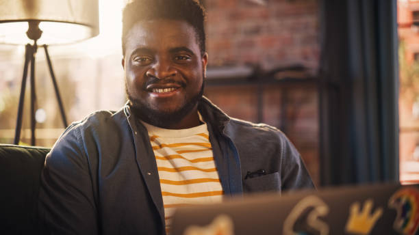 istock Handsome African American Man Working from Home on Laptop Computer in Sunny Stylish Loft Office. Creative Happy Black Male Sitting on a Couch, Smiling. Urban City View from Big Window. 1395320684