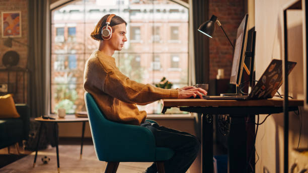 istock Young Handsome Man Working from Home on Desktop Computer in Sunny Stylish Loft Apartment. Creative Designer Wearing Cozy Yellow Sweater and Headphones. Urban City View from Big Window. 1395320723