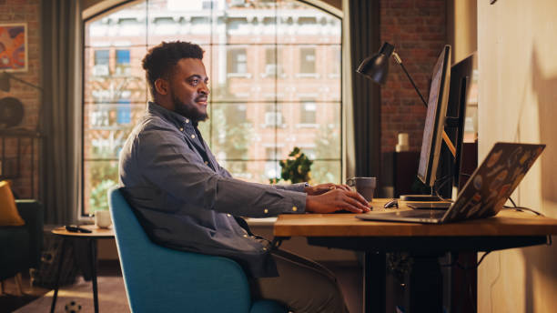 istock Young Handsome African American Man Working from Home on Desktop Computer in Sunny Stylish Loft Apartment. Creative Male Checking Social Media, Browsing Internet. Urban City View from Big Window. 1395320793