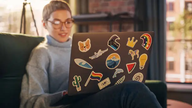 Stylish Female Using Laptop Computer with Diverse LGBT and Lifestyle Stickers on the Back. Young Creative Woman Sitting on a Couch, Typing, Browsing Internet and Checking Social Media at Home.