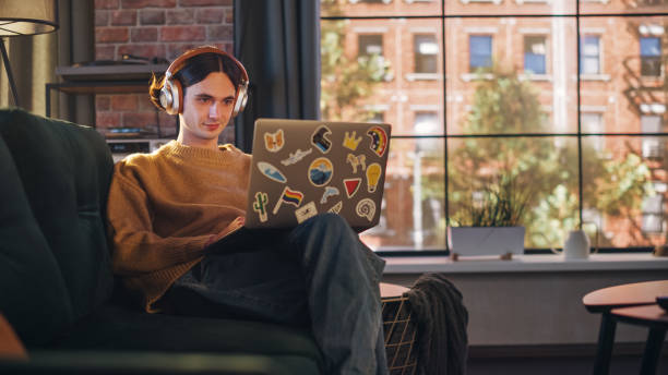 istock Young Handsome Man Sitting on Sofa and Using Laptop Computer in Sunny Stylish Loft Apartment. Creative Designer Wearing Cozy Yellow Sweater and Headphones. Urban City View from Big Window. 1395322411