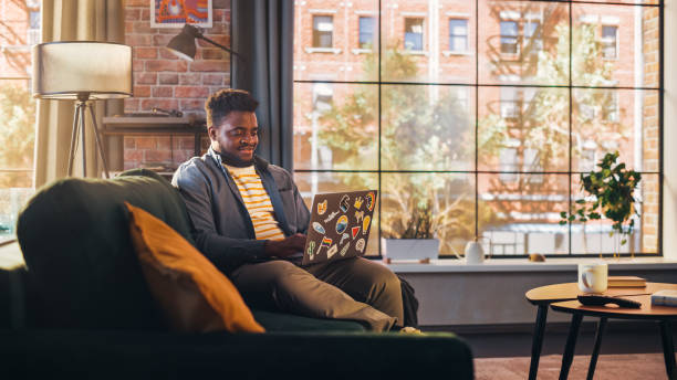 istock Young Handsome Black Man Working from Home on Laptop Computer in Sunny Stylish Loft Apartment. Creative Male Checking Social Media and Browsing Internet on a Sofa. Urban City View from Big Window. 1395322448