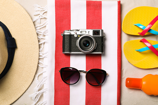 Hat, sunglasses, camera, sunscreen, beach towel and flip flops on sand, flat lay
