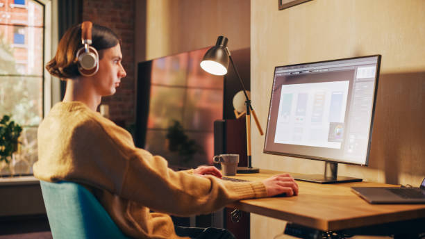istock Young Handsome Man Working from Home on Desktop Computer in Sunny Stylish Loft Apartment. Creative Designer Wearing Cozy Yellow Sweater and Headphones. Urban City View from Big Window. 1395322498