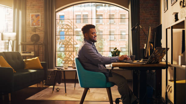 istock Young Handsome African American Man Working from Home on Desktop Computer in Sunny Stylish Loft Apartment. Creative Male Checking Social Media, Browsing Internet. Urban City View from Big Window. 1395322582