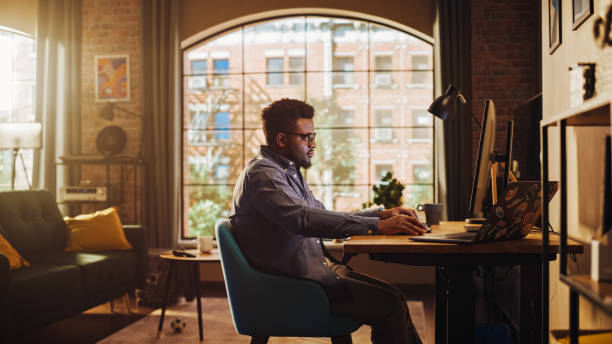 istock Young Handsome Black Man Working from Home on Desktop Computer in Sunny Stylish Loft Apartment. Creative Male Checking Social Media, Browsing Internet. Urban City View from Big Window. 1395322593