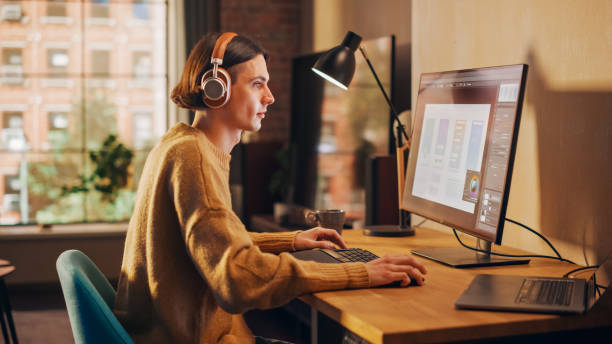 istock Young Handsome Man Working from Home on Desktop Computer in Sunny Stylish Loft Apartment. Creative Designer Wearing Cozy Yellow Sweater and Headphones. Urban City View from Big Window. Handheld Shot. 1395322633