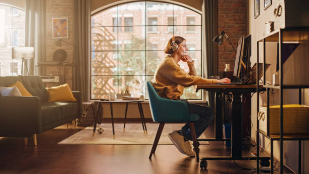 istock Young Handsome Man Working from Home on Desktop Computer in Sunny Stylish Loft Apartment. Creative Designer Wearing Cozy Yellow Sweater and Headphones. Urban City View from Big Window. 1395322653