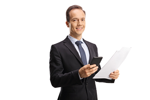 Medium shot portrait of smiling young adult businessman holding work documents while looking at the camera standing in corridor during day