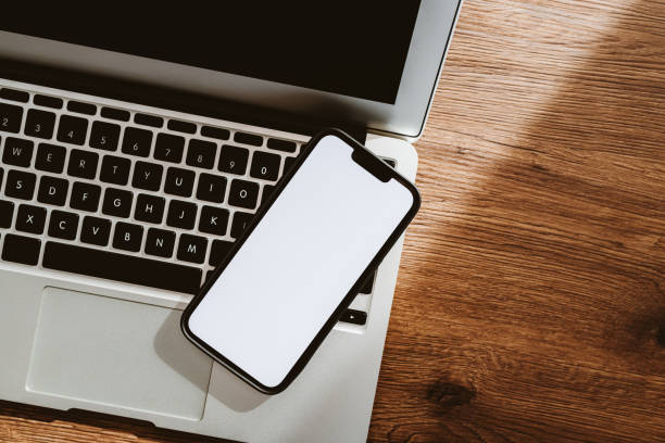 istock Mobile phone with blank white mockup touchscreen and laptop computer on office desk, top view 1395326365