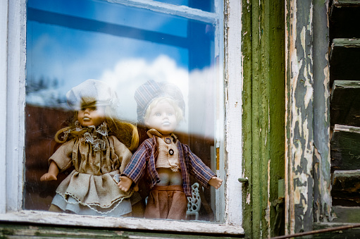 Close up color image depicting two china dolls in the window of an old house. The dolls, a girl and a boy doll, are holding hands. Room for copy space.