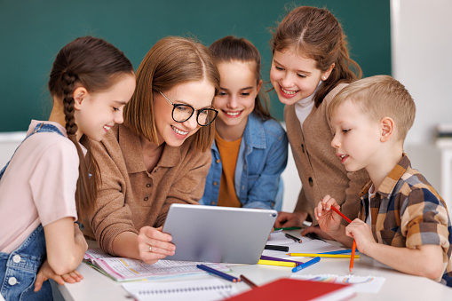 Happy female tutor and optimistic children smiling and watching video on tablet while gathering around table during lesson at school