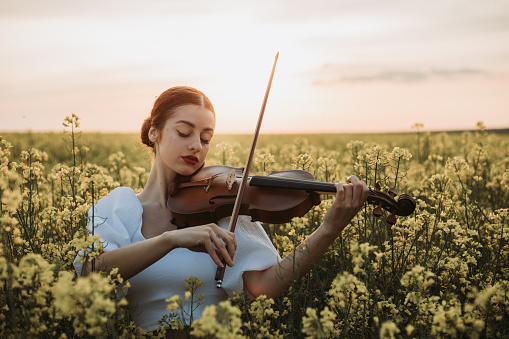 Ginger hair beautiful young woman in white dress playing violin in the flower field. Front view.