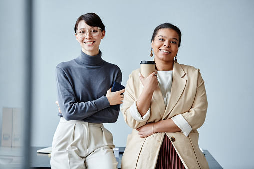 Candid waist up portrait of two young businesswomen smiling at camera in office against simple background