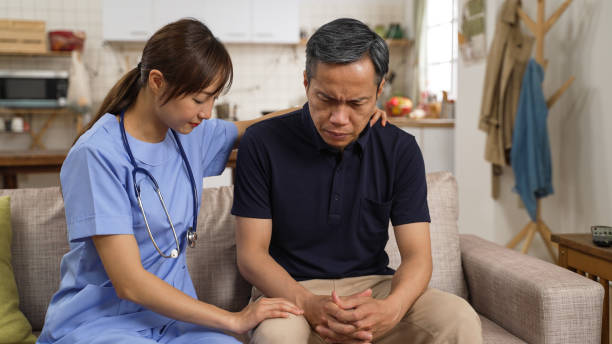 malheureux homme mûr asiatique assis avec son psychologue sur un canapé à la maison avec une expression triste. la femme attentionnée faisant preuve d’empathie et lui tapotant les genoux tout en le consolant - pat on back photos et images de collection