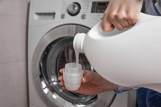 From a white plastic bottle, a rinse is poured into the cap In bathroom where washing machine is located, girl pours the laundry conditioner into lid to give clean laundry pleasant smell and softness. From white plastic bottle, rinse is poured into the cap laundry detergent stock pictures, royalty-free photos & images