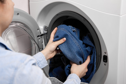 In laundry room, female person loads dirty laundry items into the washing machine. Woman in light-colored shirt pulls dirty denim out soft basket and puts them in drum of washing machine for washing.