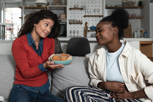 Young hungry African American woman is on diet and her friend and roommate offering to her a muffins to eat. Dieting and hunger healthy eating concept.