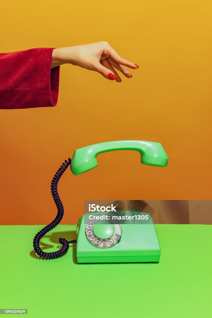 Colorful bright image of female hand holding old-fashioned green colored phone, handset falling down isolated over orange background Colorful bright image of female hand holding old-fashioned green colored phone, handset falling down isolated over orange background. Concept of pop art, vintage things, mix old and modernity Surrealism Stock Photo