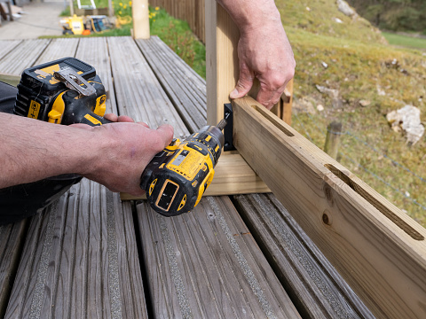 A carpenter working outdoors, building wooden decking on the side of family home. Using a handheld cordless battery drill he is securing the wooden railing posts on the edge of the deck.