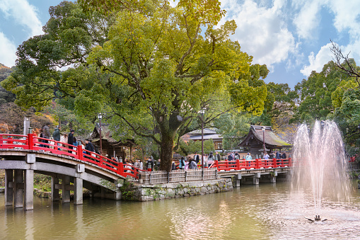 kyushu, japan - december 07 2019: Tourist crossing over the Japanese red Taiko Bashi bridge of Dazaifu Shinto Shrine above a pond with a fountain jet overlooked by large kusunoki camphor trees.