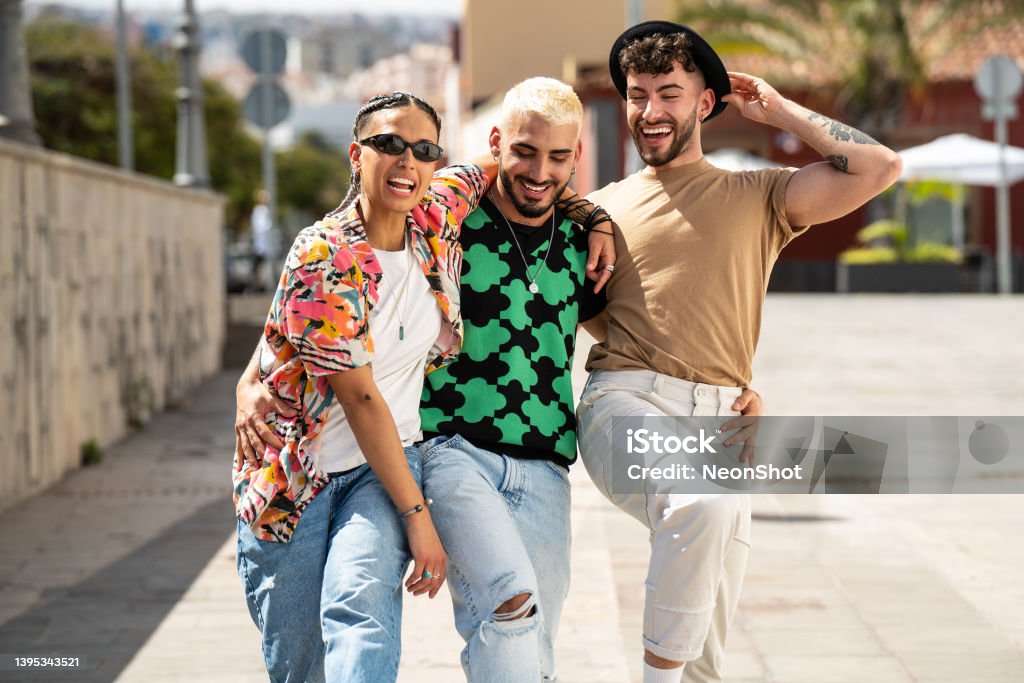 Three happy young people walking in the city, talking to each other, laughing and having fun. Multiethnic group of friends. Three happy young people walking in the city, talking to each other, laughing and having fun. Multiethnic group of friends. Real people lifestyle. LGBTQIA People Stock Photo