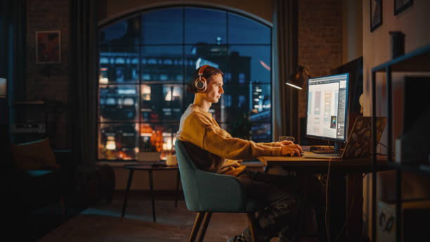 istock Young Handsome Man Working from Home on Desktop Computer in Stylish Loft Apartment in the Evening. Creative Designer Wearing Cozy Yellow Sweater and Headphones. Urban City View from Big Window. 1395344474