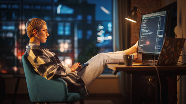 istock Young Woman Writing Code on Computer in Stylish Loft Apartment in the Evening. Creative Female Wearing Cozy Clothes, Resting Feet on Table and Using Wireless Keyboard. Urban City View from Big Window. 1395344798