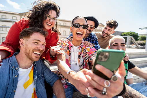 Group of happy multiracial friends using smartphone outdoors, watching social media and laughing. Fashionable people making a selfie.
