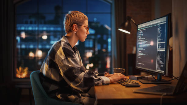 Young Woman Writing Code on Desktop Computer in Stylish Loft Apartment in the Evening. Creative Female Wearing Cozy Clothes, Working from Home on Software Development. Urban City View from Big Window. mujer joven escribiendo código en una computadora de escritorio en un elegante apartamento loft por la noche. mujer creativa con ropa acogedora, trabajando desde casa en el desarrollo de software. vista urbana de la ciudad desde una gran ventana.