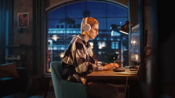 Young Woman Writing Code on Desktop Computer in Stylish Loft Apartment in the Evening. Creative Female Wearing Headphones, Working from Home on Software Development. Urban City View from Big Window.