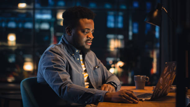 istock Young Handsome Black Man Working from Home on Laptop Computer in Stylish Loft Apartment in the Evening. Creative Male Gets a Great Idea while Checking Social Media. Urban City View from Big Window. 1395344929