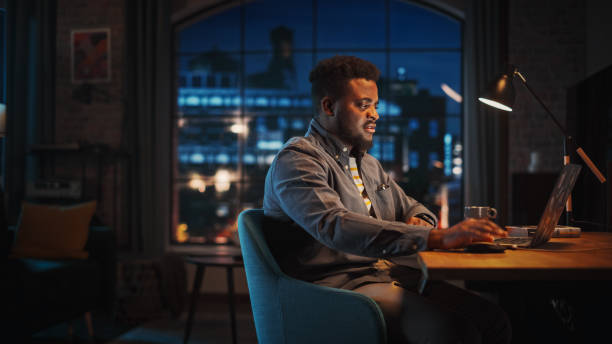 istock Young Handsome African American Man Working from Home on Laptop Computer in Stylish Loft Apartment at Night. Creative Male Checking Social Media, Browsing Internet. Urban City View from Big Window. 1395344941