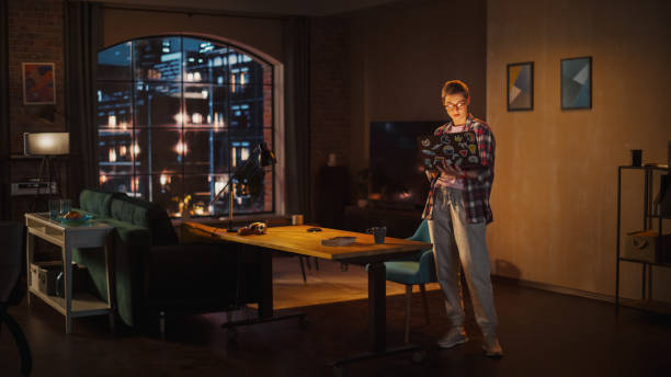 istock Young Beuatiful Women Working from Home, Standing And Holding Laptop Computer in Stylish Loft Apartment in the Evening. Creative Female Wearing Cozy Casual Outfit. Urban City View from Big Window. 1395344976