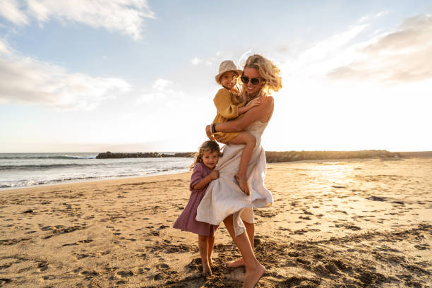 felice bella famiglia che si rilassa e cammina sulla spiaggia del tramonto in estate. mamma sorridente con le sue due figlie piccole che si divertono in vacanza. - beach women joy sand foto e immagini stock