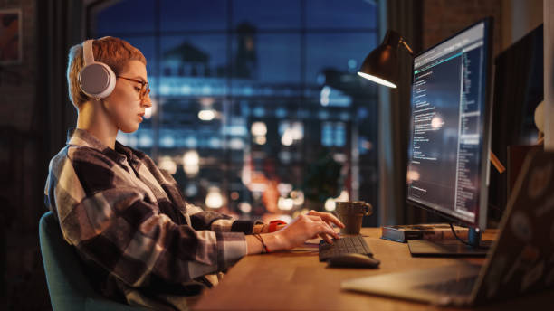 istock Young Woman Writing Code on Desktop Computer in Stylish Loft Apartment in the Evening. Creative Female Wearing Headphones, Working from Home on Software Development. Urban City View from Big Window. 1395345277
