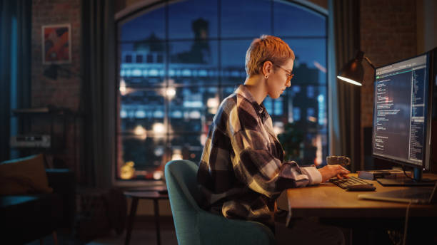 istock Young Woman Writing Code on Desktop Computer in Stylish Loft Apartment in the Evening. Creative Female Wearing Cozy Clothes, Working from Home on Software Development. Urban City View from Big Window. 1395345314