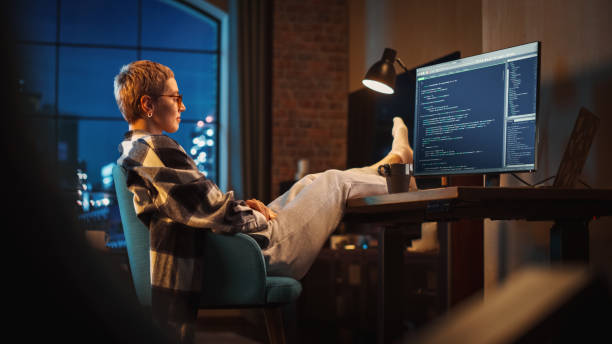 istock Young Woman Writing Code on Computer in Stylish Loft Apartment in the Evening. Creative Female Wearing Cozy Clothes, Resting Feet on Table and Using Wireless Keyboard. Urban City View from Big Window. 1395345336