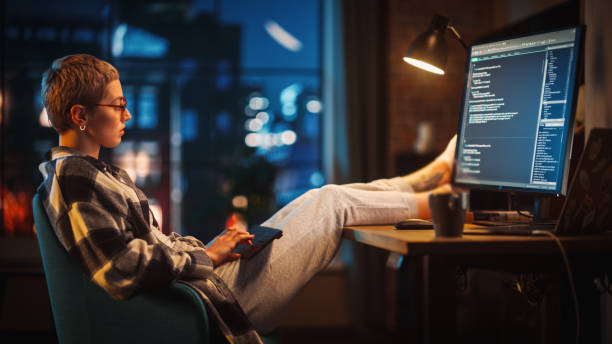 istock Young Woman Writing Code on Computer in Stylish Loft Apartment in the Evening. Creative Female Wearing Cozy Clothes, Resting Feet on Table and Using Wireless Keyboard. Urban City View from Big Window. 1395345354
