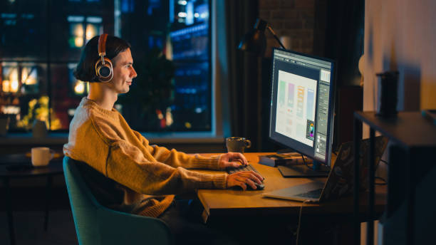 istock Young Handsome Man Working from Home on Desktop Computer in Stylish Loft Apartment in the Evening. Creative Designer Wearing Cozy Yellow Sweater and Headphones. Urban City View from Big Window. 1395345968