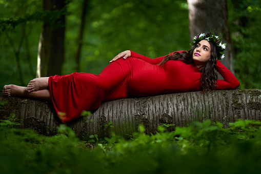 Portrait of a beautiful young hispanic pregnant woman in long red dress in a deciduous forest in the summer
