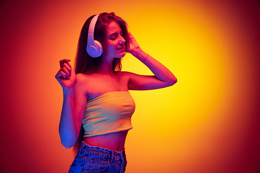 Looks delightful. Portrait of young girl listening to music in headphones isolated over gradient red yellow background in neon. Concept of beauty, art, fashion, youth and emotions. Copy space for ad
