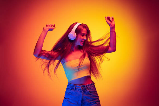 Dynamic portrait of young beautiful girl listening to music in headphones and dancing isolated over gradient red yellow background in neon stock photo