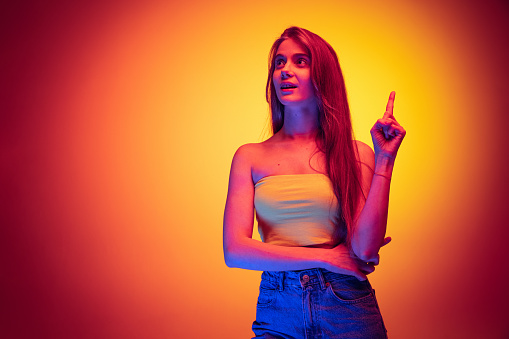 Thoughtful. Portrait of young woman in casual cloth rising finger up with idea isolated over gradient red yellow background in neon light. Concept of beauty, art, fashion, youth and emotions.