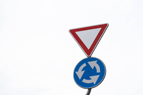 A prominent yellow diamond-shaped warning sign stands out at an intersection surrounded by trees and traffic signals on a cloudy day.