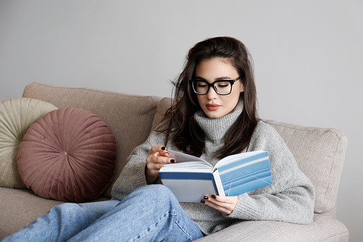 Young beautiful woman wearing grey turtleneck reading a book at home. Brunette female in a sweater sitting on beige textile couch in her lofty apartment. Background, copy space, close up.